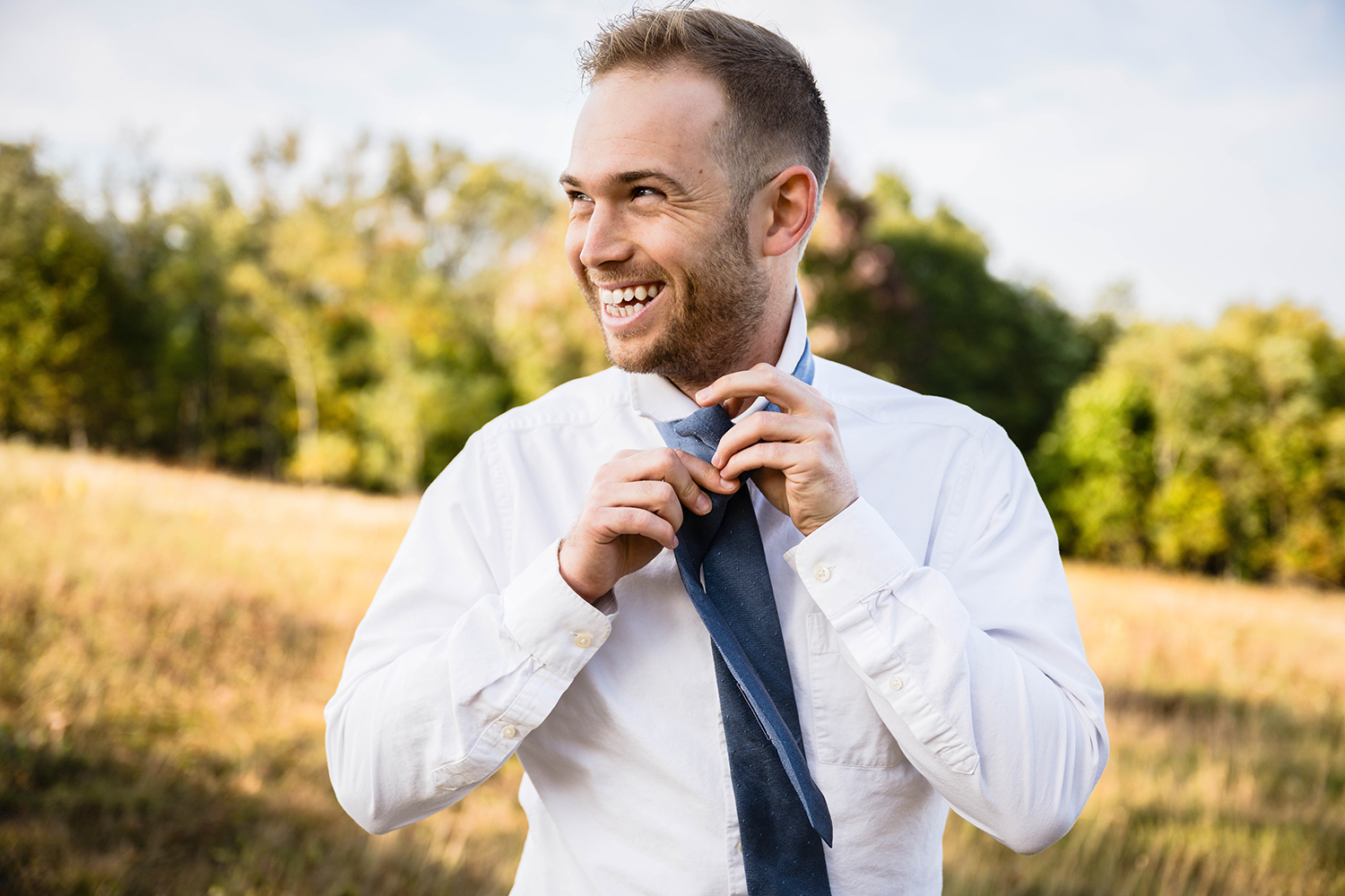 A groom ties his tie as he looks at his bride off camera with a big smile on his face.