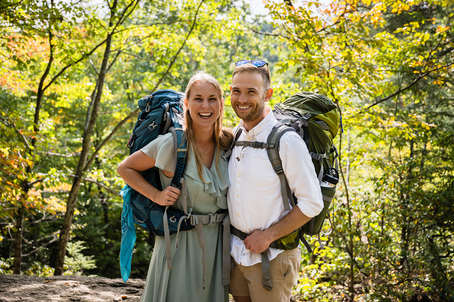 A bride and groom smile widely with backpacks on their backs as they take a moment to pause and grab a photo together during their hike on the Dark Hallow Falls trail in Shenandoah National Park. 