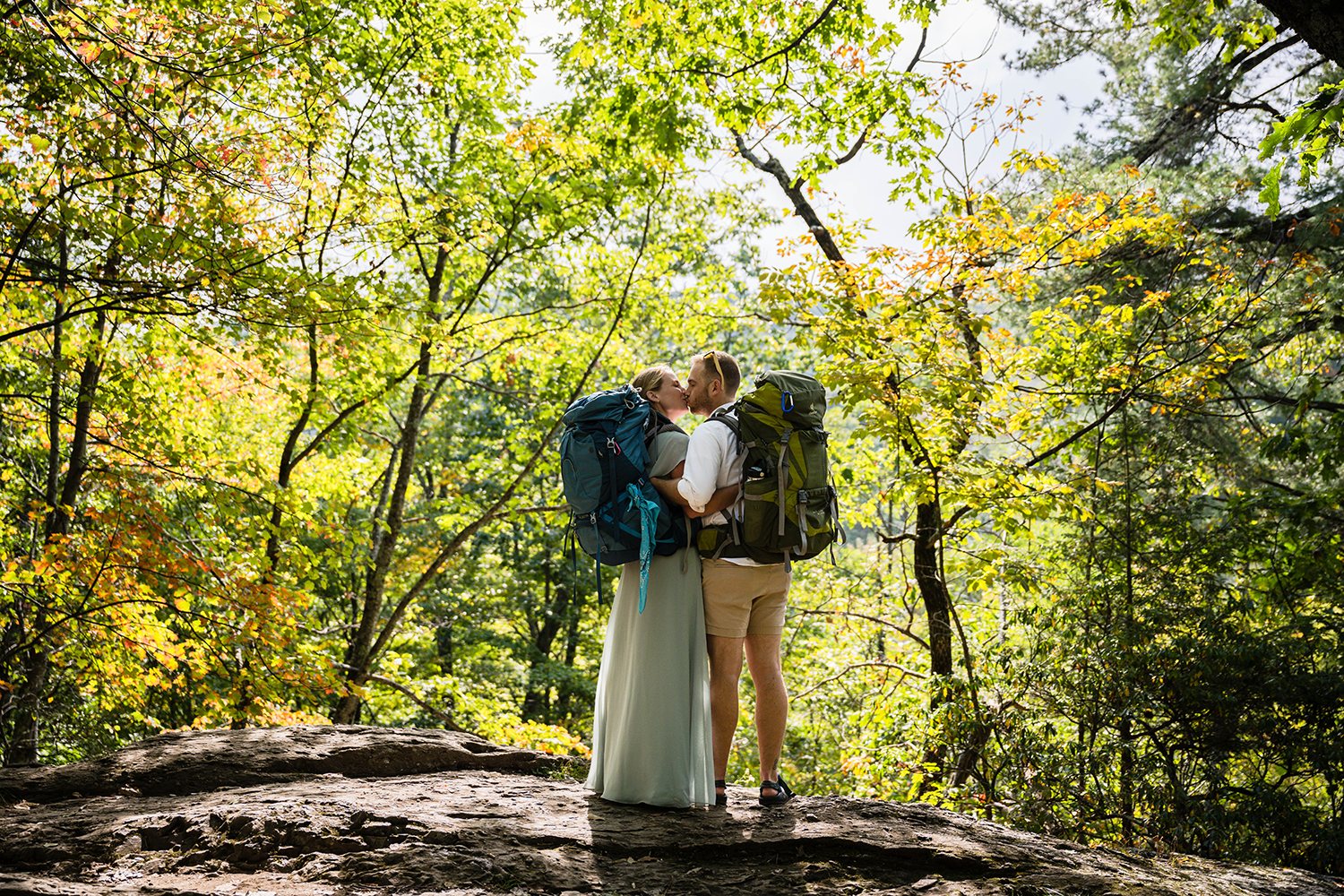 A couple wearing large backpacks hold onto one another and kiss while standing on a rock a little off the trail.