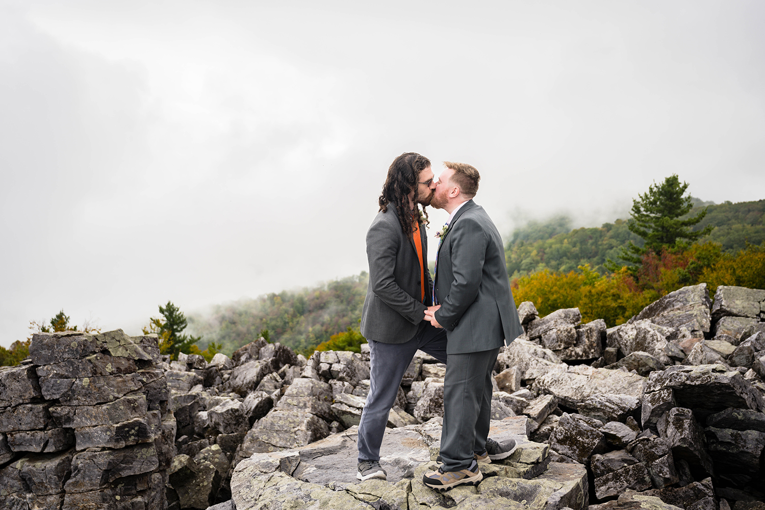 An LGBTQ+ couple stand in front of one another kissing as they hold their partner's hands in front of them while standing on a large rock at Blackrock Summit in Shenandoah National Park.