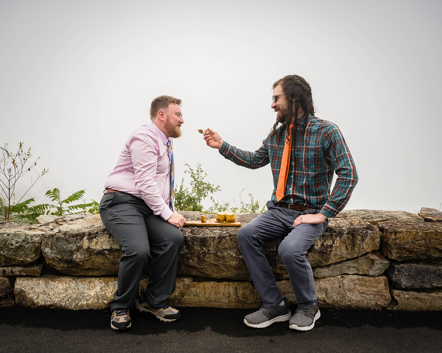 Two gay marriers sit on a stone guardrail at Loft Mountain Overlook in Shenandoah National Park to cut their wedding cake and feed pieces of it to one another.