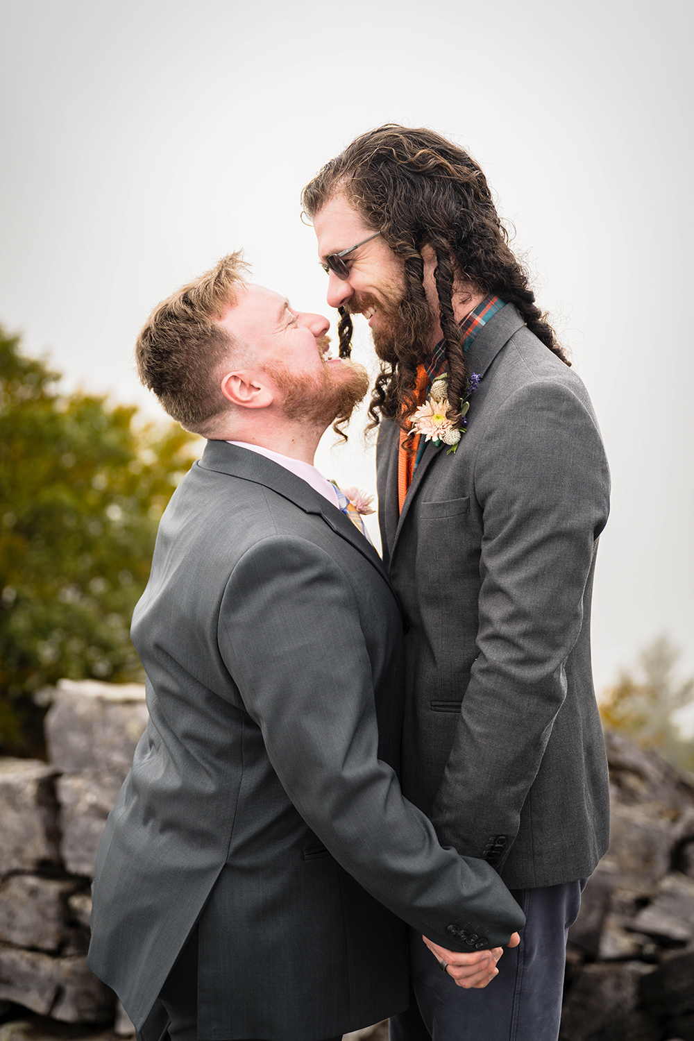 A queer couple smiles widely showing their teeth as they got in for a kiss and holding hands at Blackrock Summit.