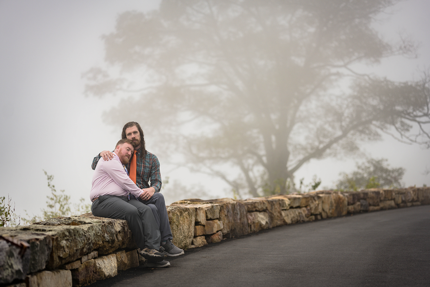 An LGBTQ+ couple sits closely on a stone wall, enveloped in a serene fog, at Loft Mountain Overlook.