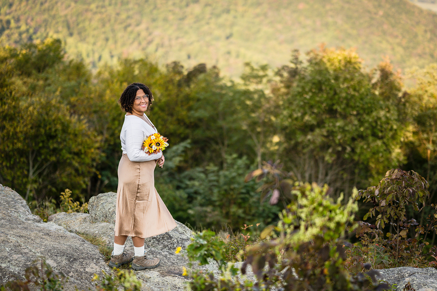 A marrier holding a DIY bouquet of fake flowers looks over her shoulder for a photo as she stands on a rock at Timber Hollow Overlook in Shenandoah National Park.