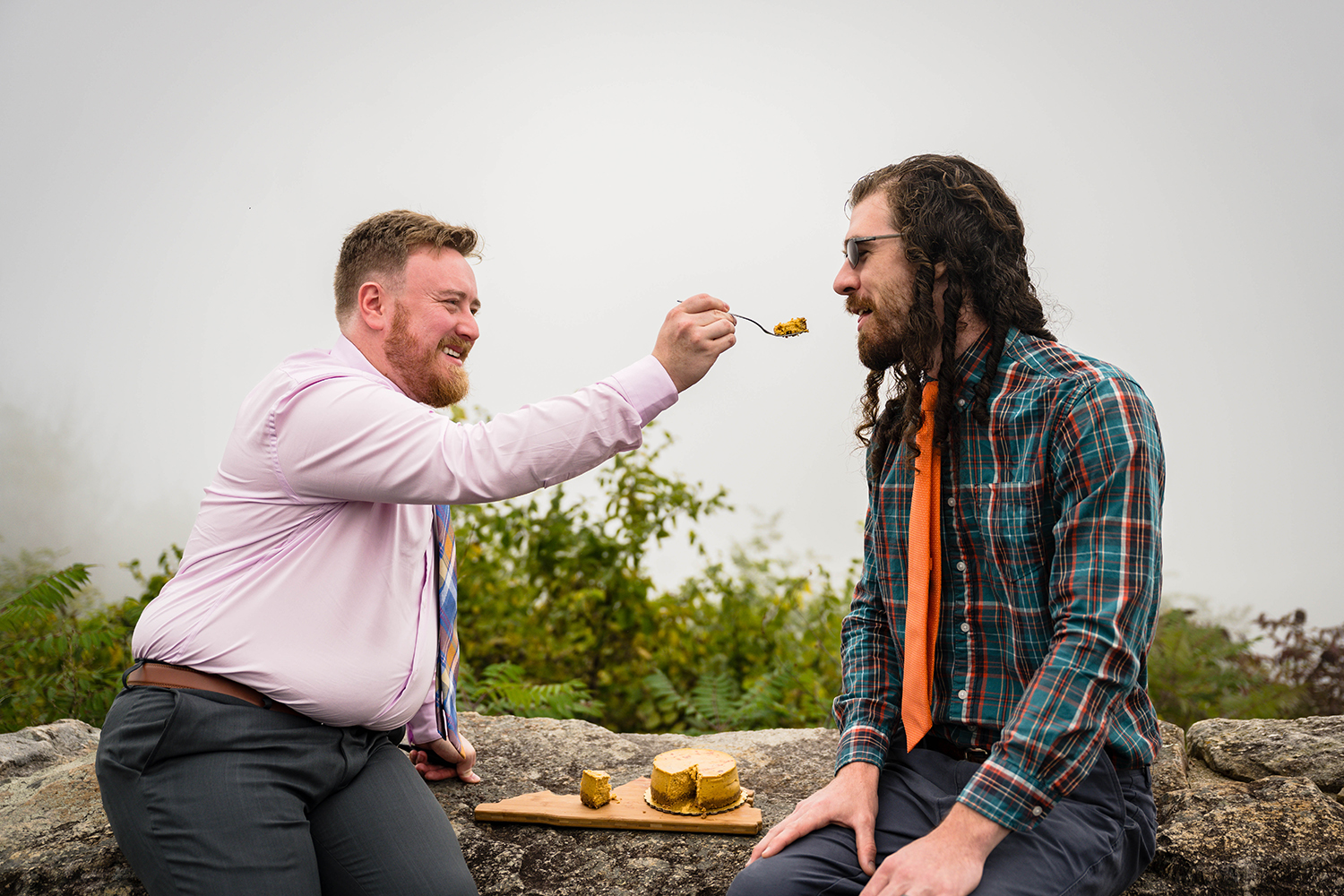 Two LGBTQ+ marriers sit on a stone guardrail at Loft Mountain Overlook in Shenandoah National Park to cut their wedding cake and feed pieces of it to one another.