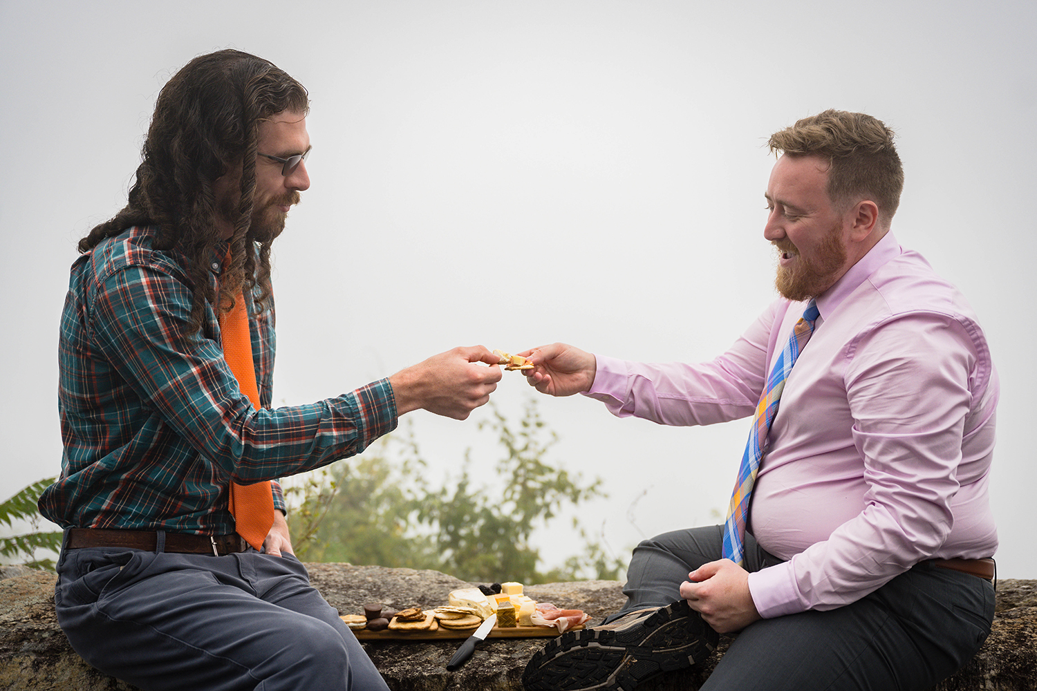 Two queer marriers sitting on a stone guardrail outdoors, dressed in business casual attire. They are sharing food, with both marriers holding onto crackers stacked with meats and cheeses. 