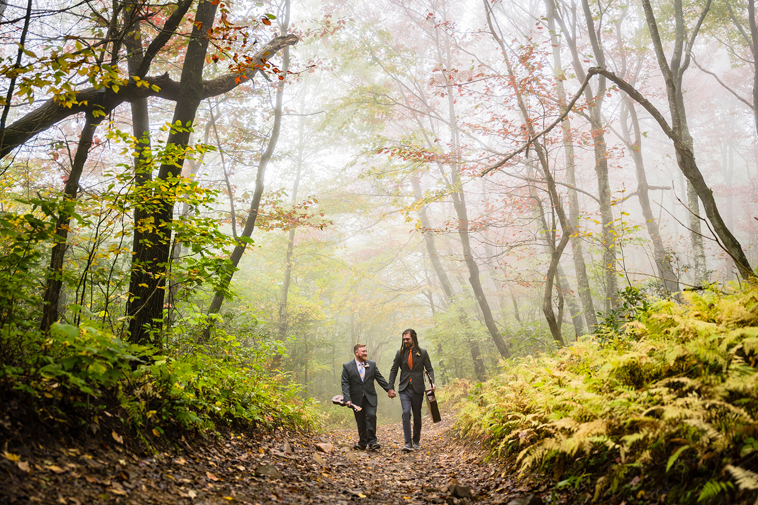 A gay couple walk hand-in-hand in a foggy forest with vibrant green, orange, and red leaves while holding onto their instruments in their free hands.