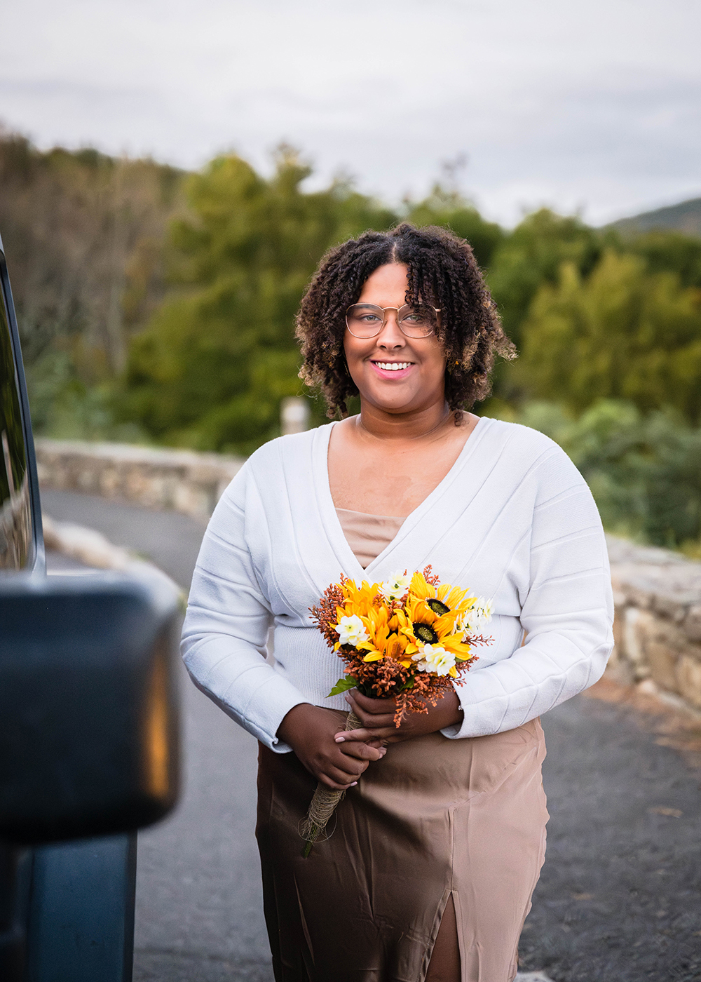 A bride holds a DIY bouquet of flowers and smiles for a photo as she stands next to her Jeep.