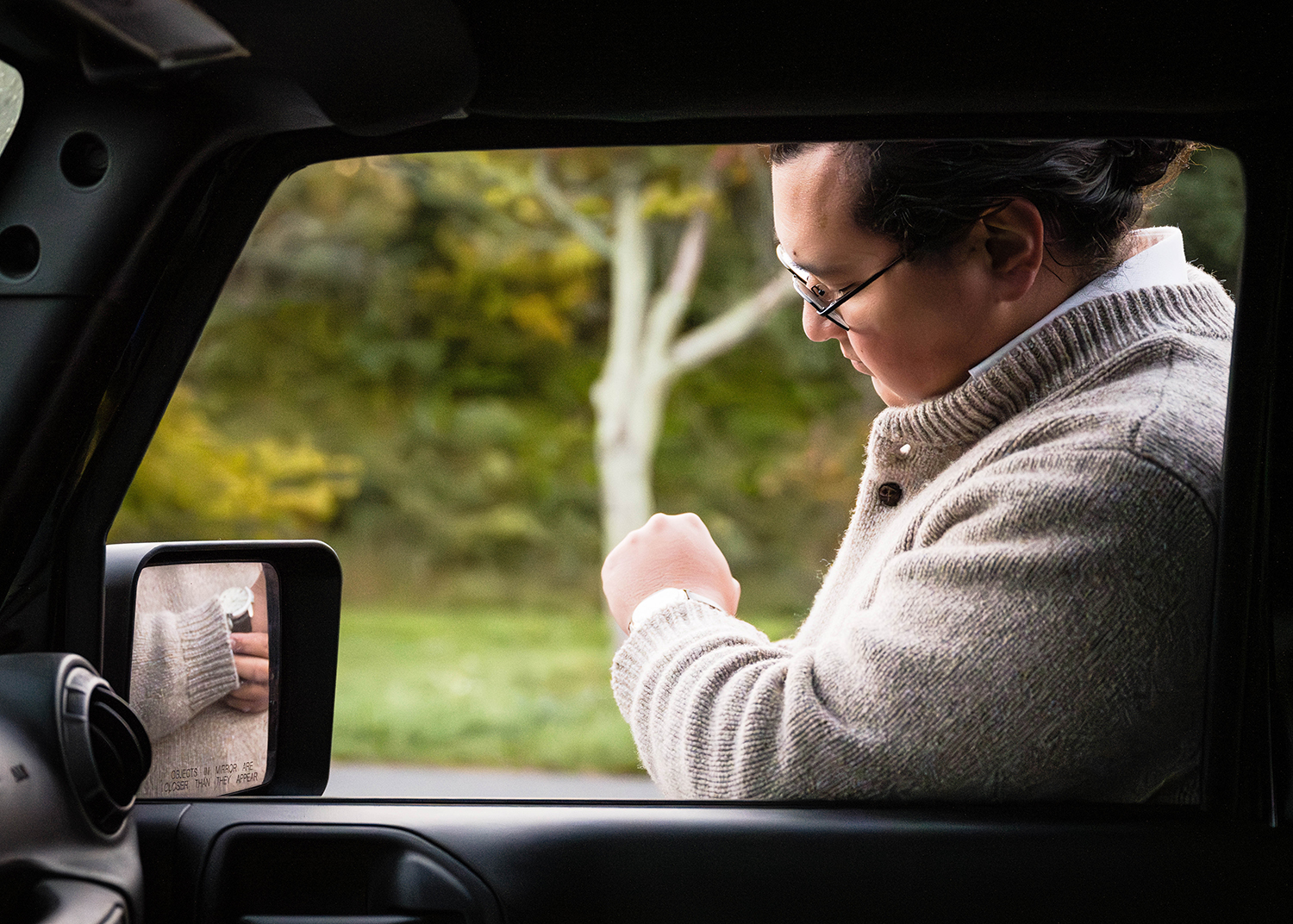 A groom looks down as he adjusts his watch. He uses the rearview mirror outside of his Jeep to make the adjustments. 