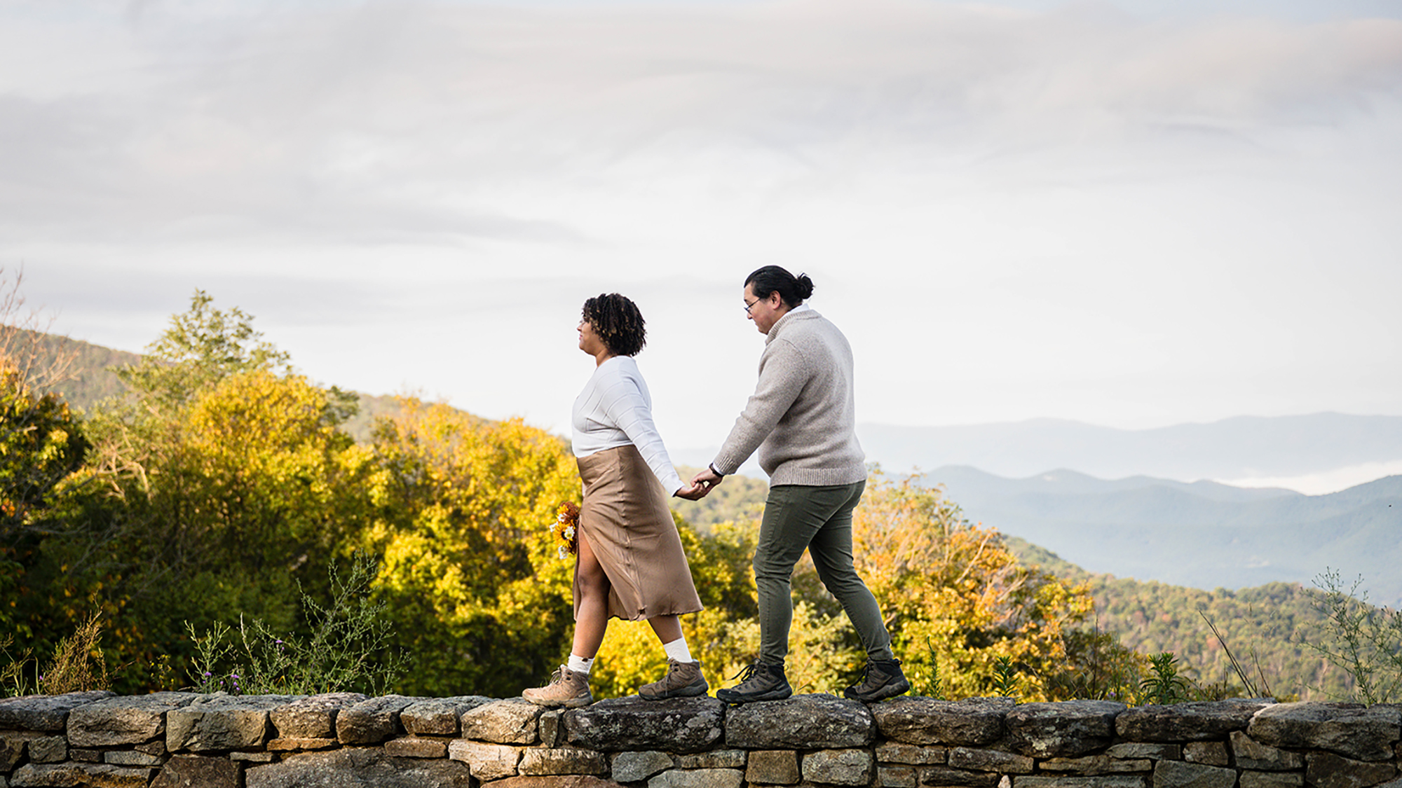 A couple walk hand-in-hand along a stone wall at Timber Hollow Overlook in Shenandoah National Park on their elopement day.