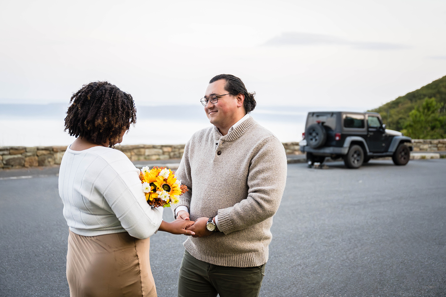 A couple on their elopement day smile softly at one another as they do their first look at Timber Hollow Overlook in Shenandoah National Park. Their Jeep is parked behind them along the curb at Timber Hollow Overlook.