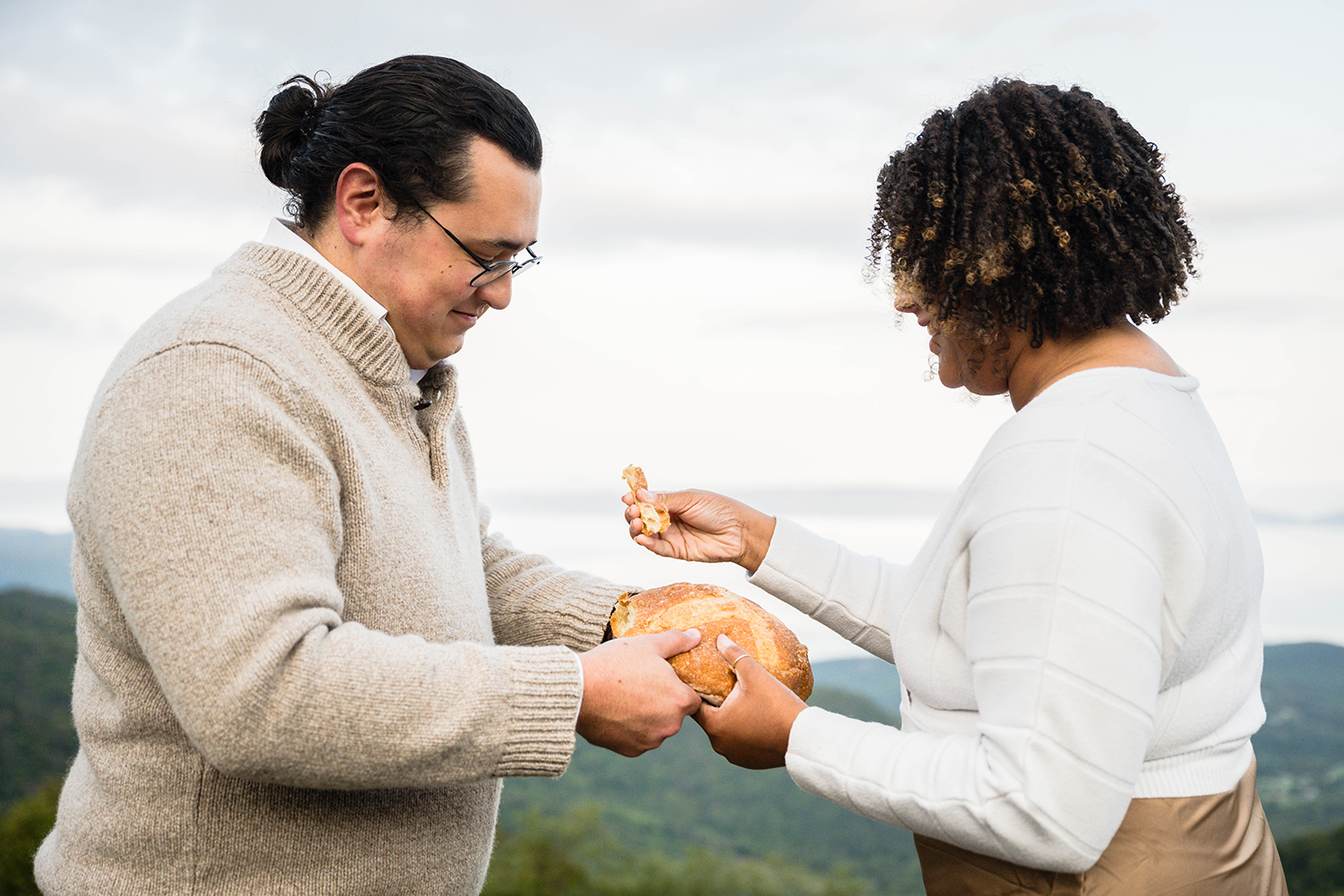 A wedding couple on their elopement day breaks a loaf of bread and begins to feed pieces of it to one another.