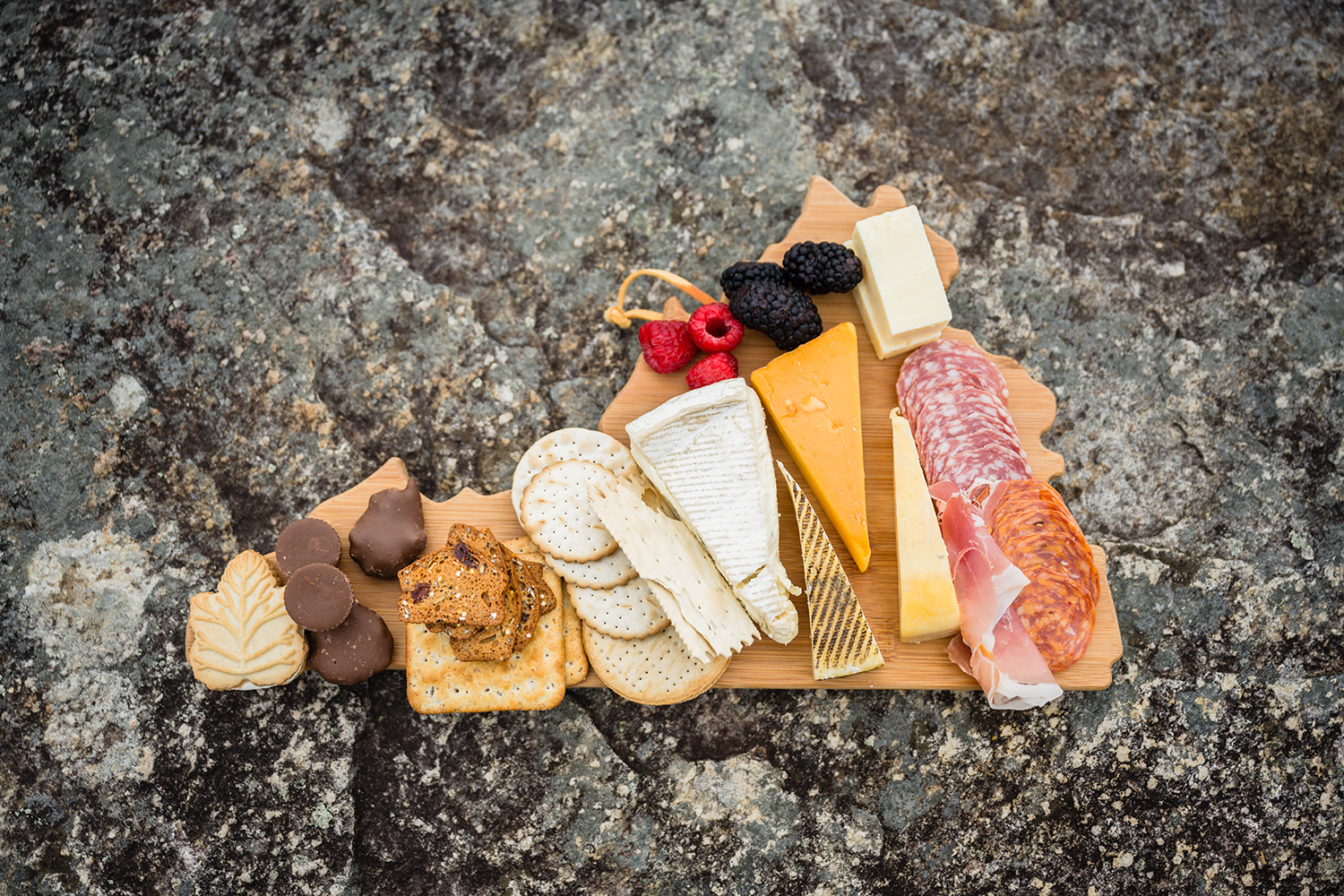 A Virginia state shaped charcuterie board with a variety of cheeses, meats, crackers, fruits, chocolates, and cookies on a stone guardrail in Shenandoah National Park.