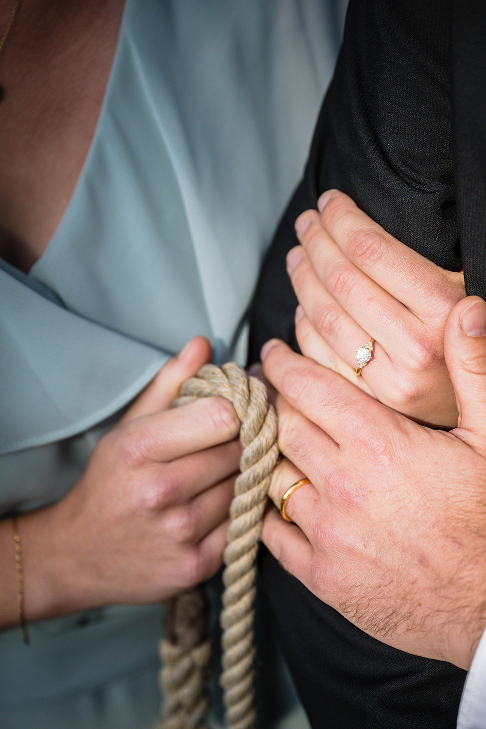 A bride wraps her hand around the groom's arm while the groom places his hand on the brides hand. Their hands both show their wedding rings. The bride also holds a tan, braided leash against the arm of the groom.