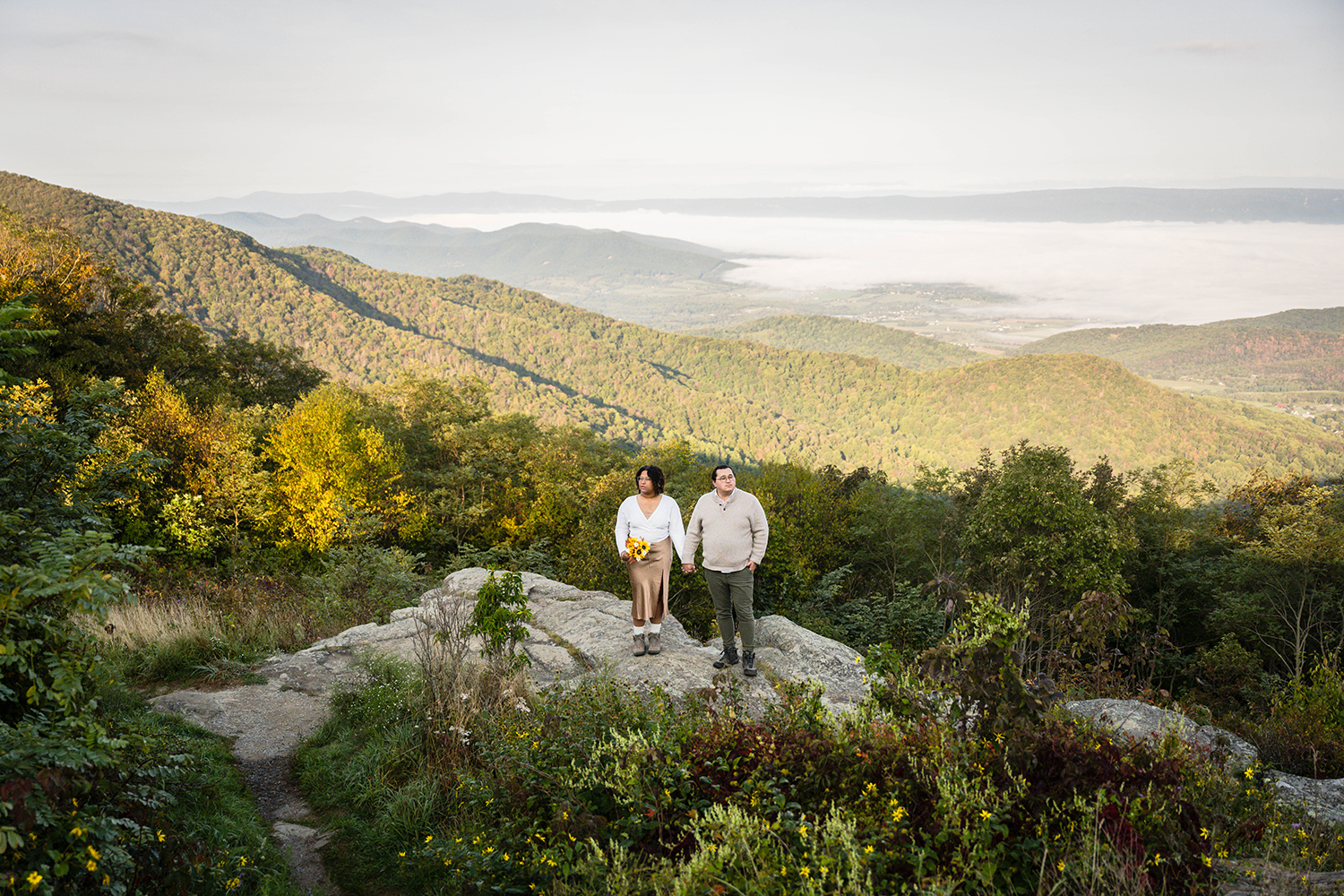 An elopement couple stands on a rock, gazing at the majestic mountains in the distance, enjoying a scenic view together at Timber Hollow Overlook in Shenandoah National Park.