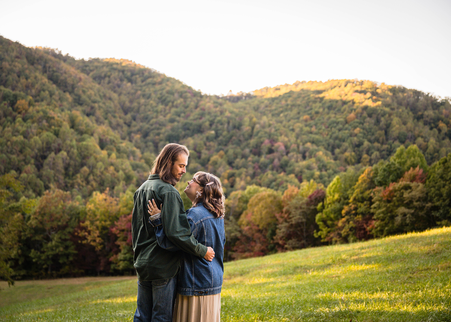 A couple wrap their arms around each other's backs and look at one another while smiling. In the background, there are rolling hills of fall colored trees.