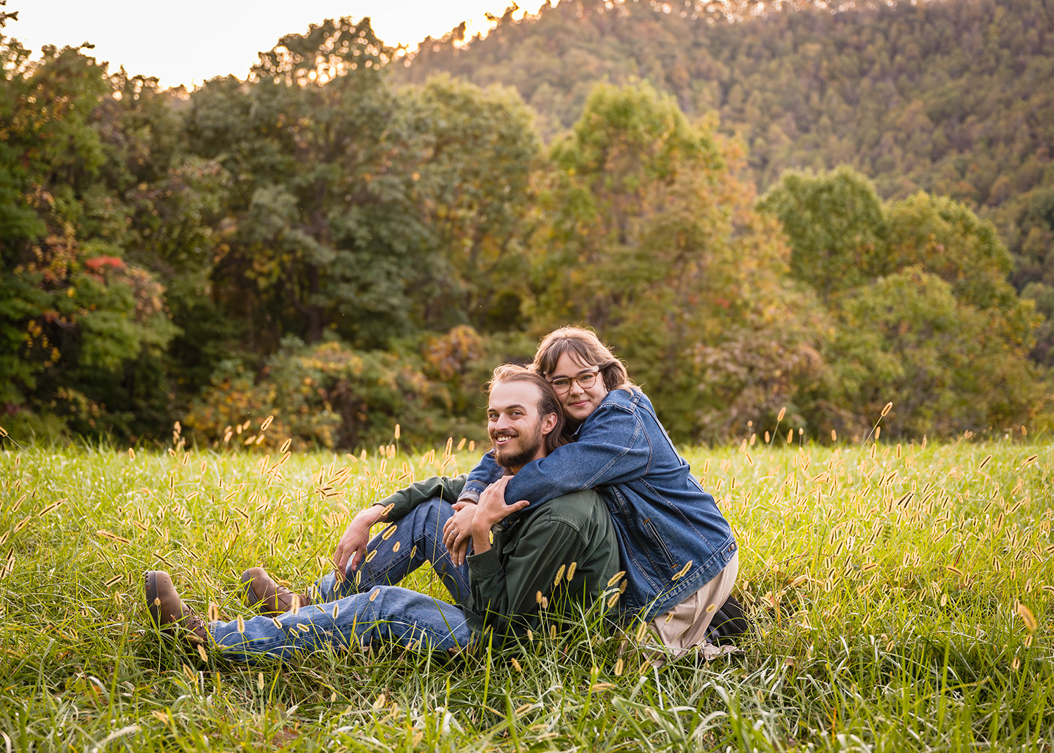 A man sits in a field on a mountain and smiles at the camera while a woman kneels behind him, wraps her arms around his neck, leans her head against his head, and smiles softly at the camera during their fall engagement session.