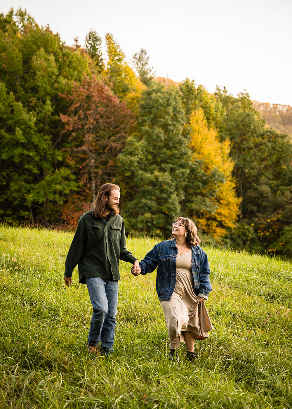 A couple walks while holding hands down a mountain with colorful trees in the background during their fall engagement adventure session in Virginia.