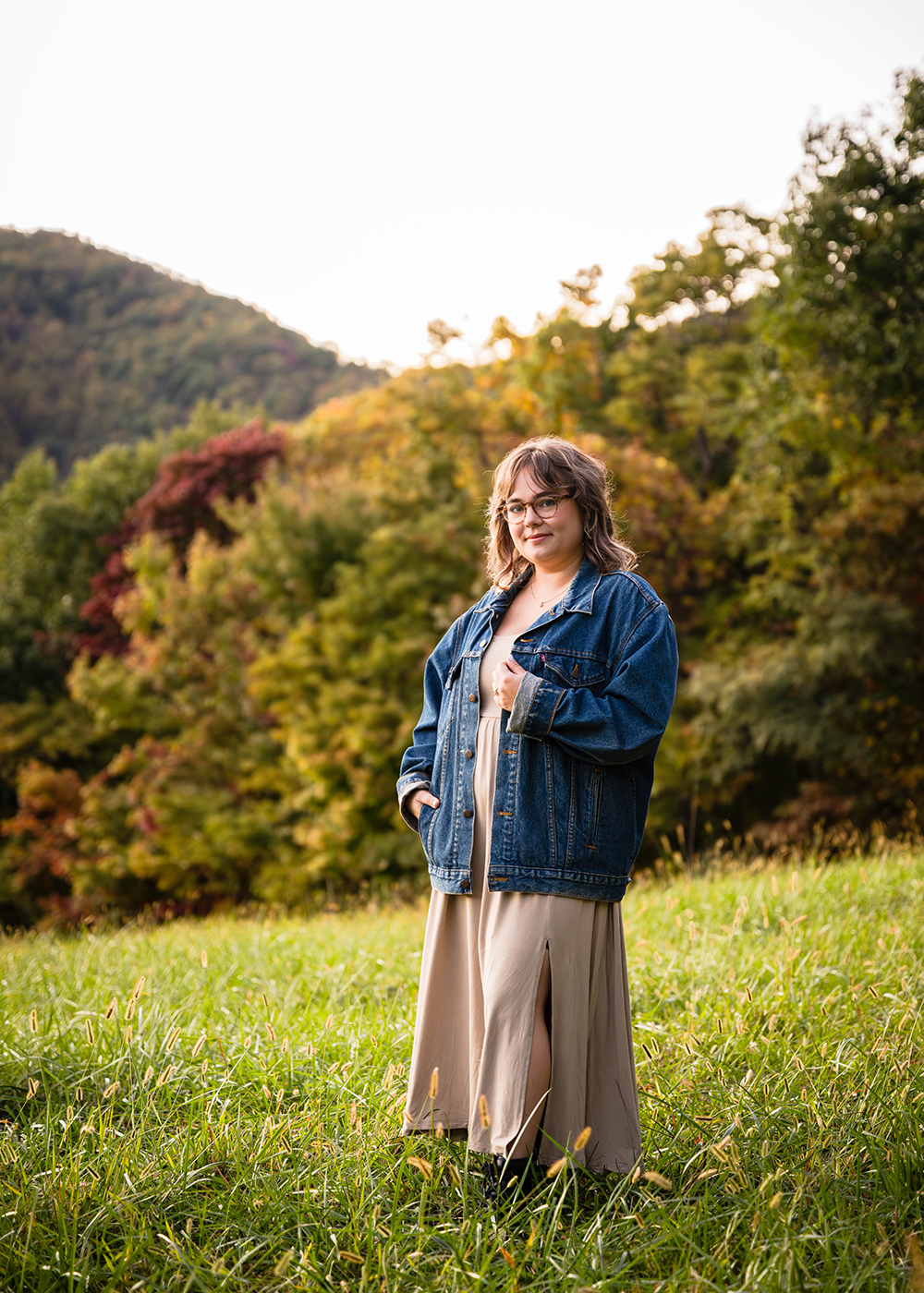 A woman during her fall engagement session poses for an individual photo with the colorful trees behind her. She is earing glasses, an oversized denim jacket, a champagne dress with a leg slit, and a pair of black Doc Marten's. 