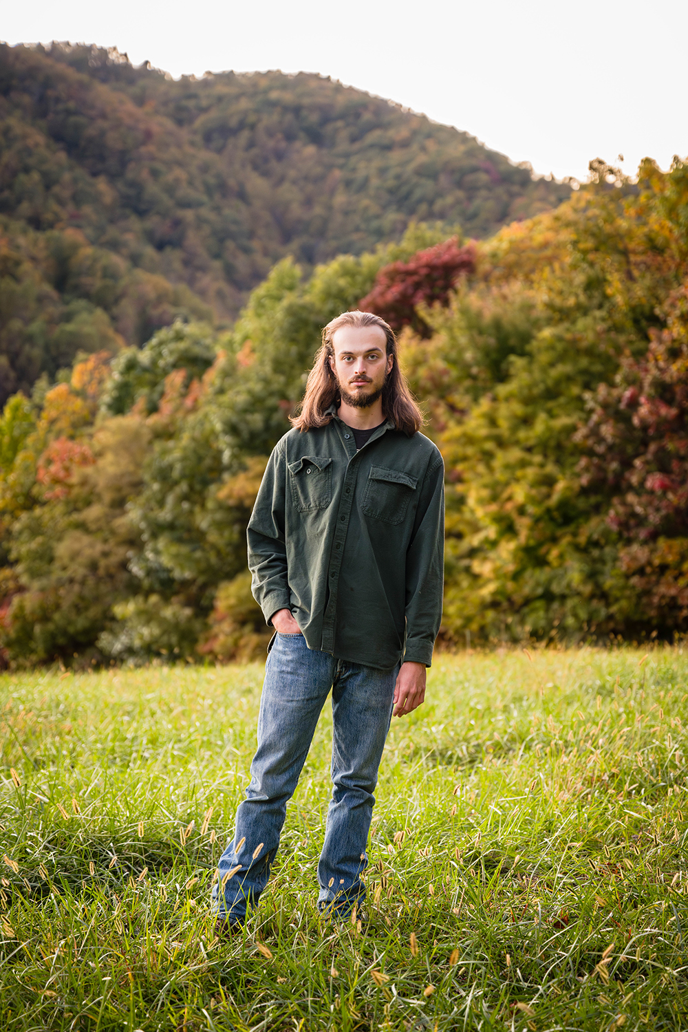 A man during his fall engagement session poses for an individual photo with the colorful trees behind him. He is wearing a green button up flannel shirt with a pair of blue jeans and his work boots.