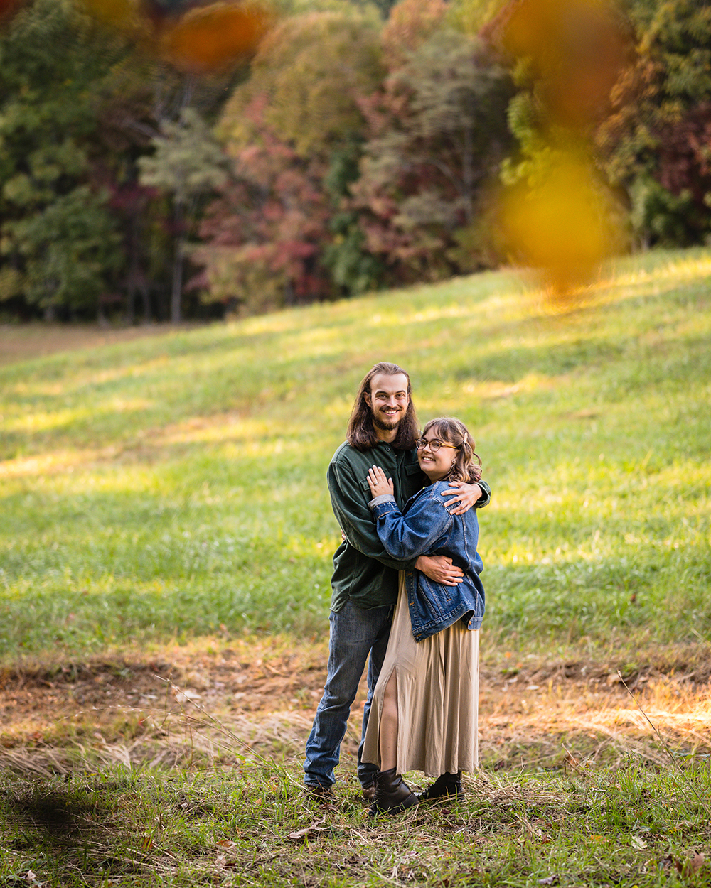 A couple during their fall engagement photoshoot smile for the camera as they pose together on a mountain. The woman wraps one arm around the man's back and places her other hand, showing her engagement ring, on the man's chest. The man wraps his arms around the woman's upper back and waist.