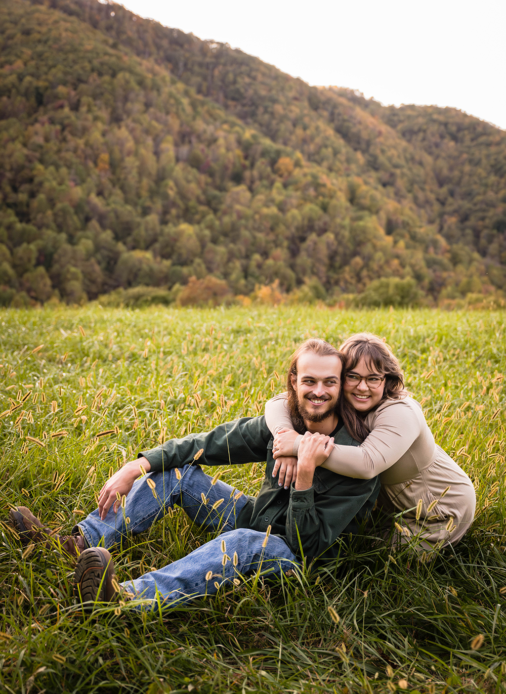 A man sits in a field on a mountain and looks away while smiling from the camera while a woman kneels behind him, wraps her arms around his neck, leans her head against his head, and also smiling and looking away from the camera during their fall engagement session.