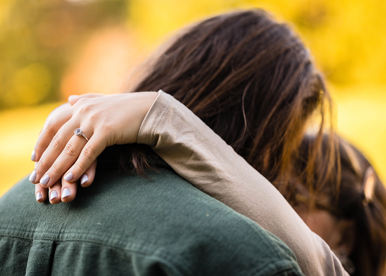 A woman during her fall engagement session wraps her arms around her partner's neck and places her hands on top of one another to reveal her engagement ring.