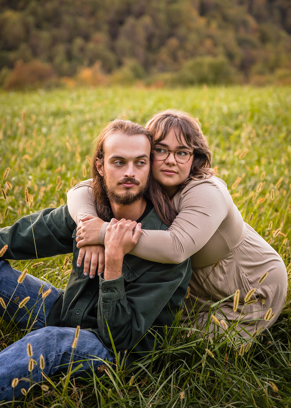 A man sits in a field on a mountain and looks away from the camera while a woman kneels behind him, wraps her arms around his neck, leans her head against his head, and also looks away from the camera during their fall engagement session.