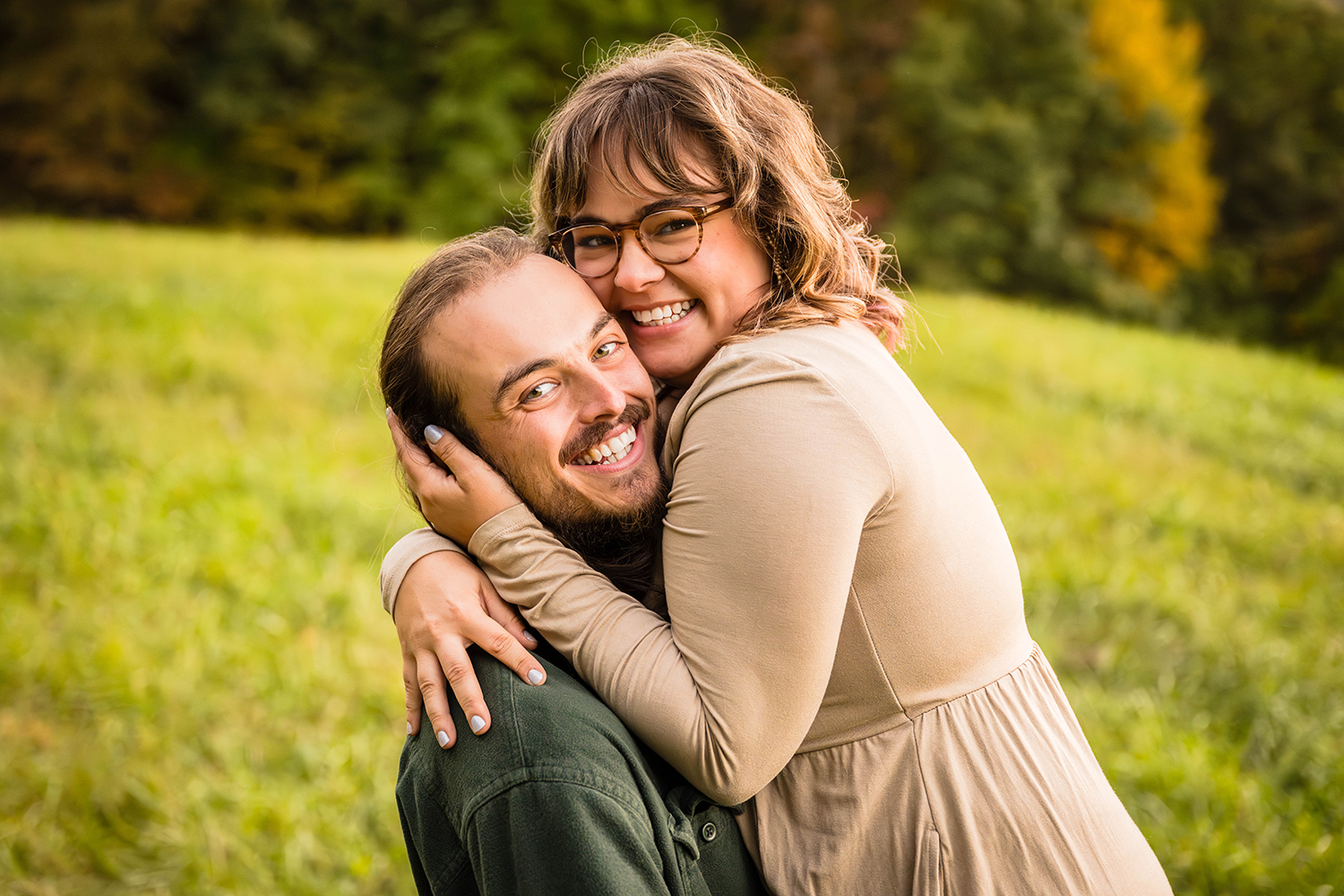 A woman holds onto her partner while he holds up during their fall engagement session. The couple smiles widely and press their heads together as the woman holds onto her partner while he holds her.