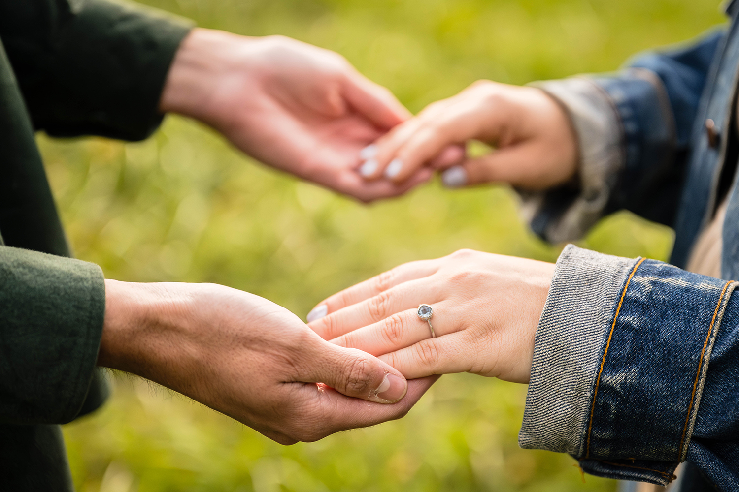 A woman places her hands into her partner's hands during their fall engagement session. The hand closest to the front shows a silver engagement ring with a blue stone.