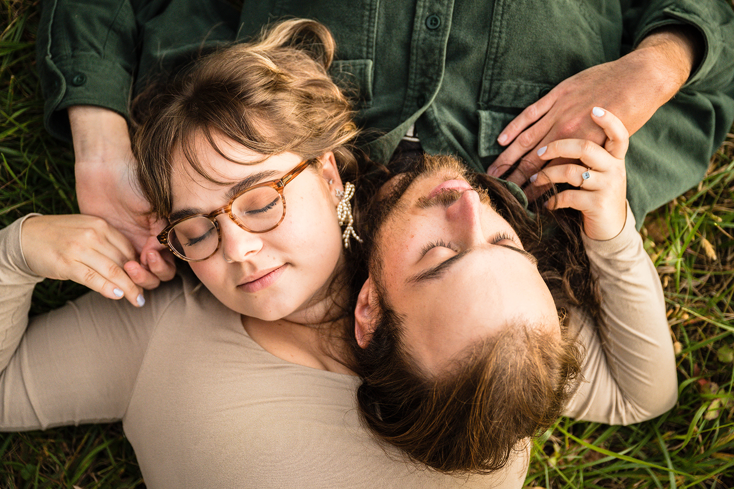 A man and woman lay on the ground in opposite directions so that they can lay their heads on top of the other's chest. They grab onto each other's hands and close their eyes for a photo.