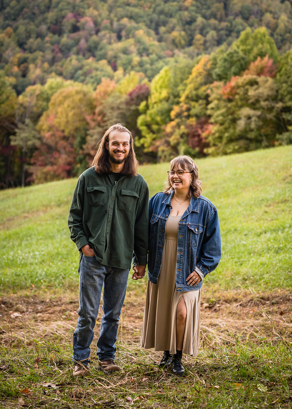 A couple during their fall engagement photo session holds hands and smiles widely for a photo. The trees behind them begin to show their changing autumn colors.