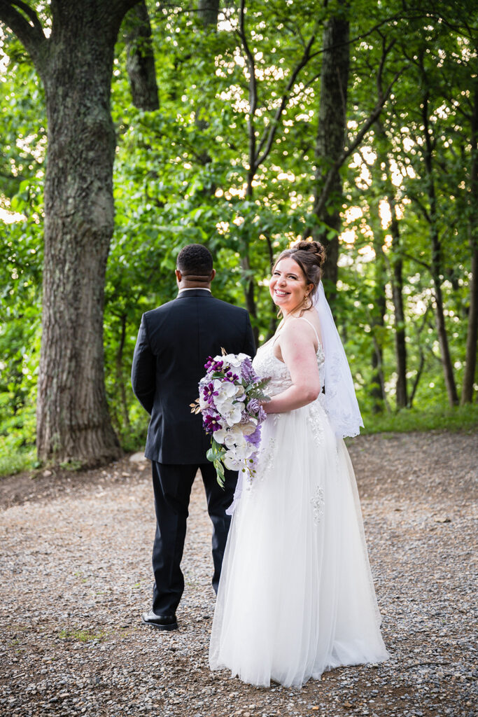 A woman smiles widely as she holds her bouquet and waits for her first look with her soon-to-be husband, who is standing in front of her with his back towards her.