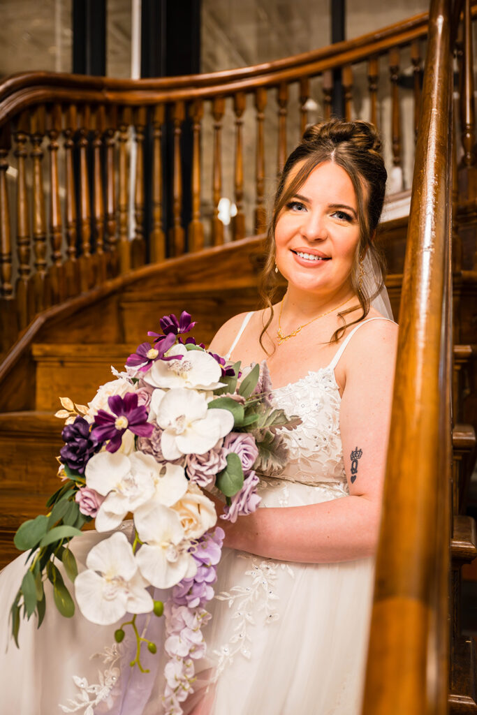 A woman sits on a stairwell and holds her wedding bouquet in her lap for a photo.