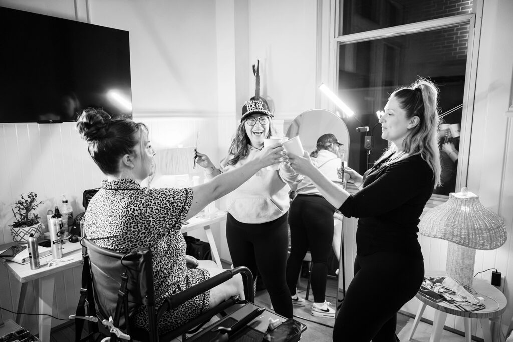 A bride, her makeup artist, and hairstylist all clink their paper cups together in a hotel room to celebrate the bride's wedding day in Roanoke, Virginia.