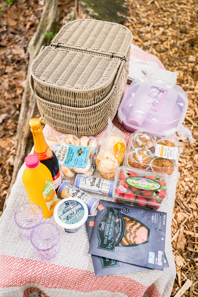 A small bench with a picnic blanket and an assortment of breakfast foods like strawberries, muffins, bagels, lox, cream cheese and breakfast drinks like cold brew and tangerine juice.