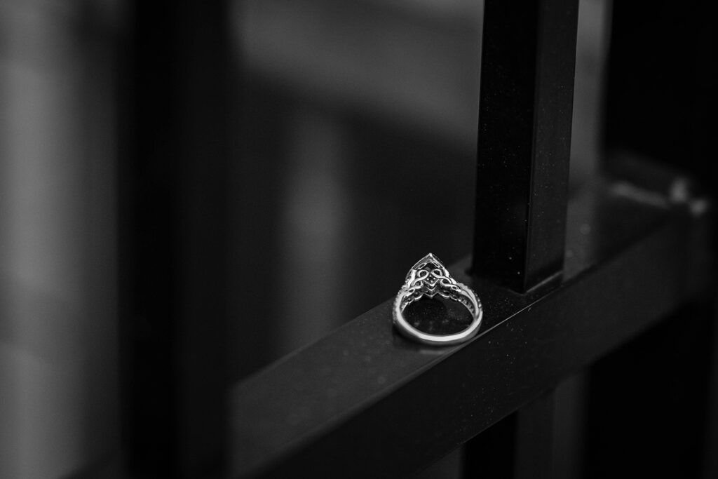 An engagement ring with intricate details is set on a fence for a photo.