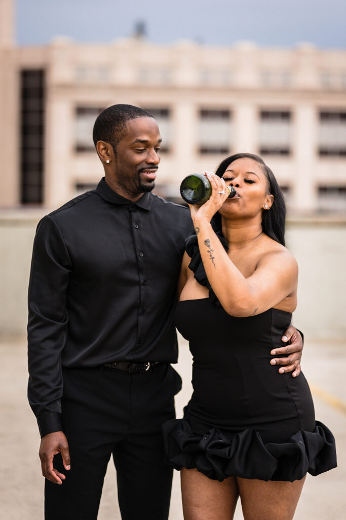 A Black woman drinks champagne directly from the bottle with her fiancé next to her during their engagement photos.