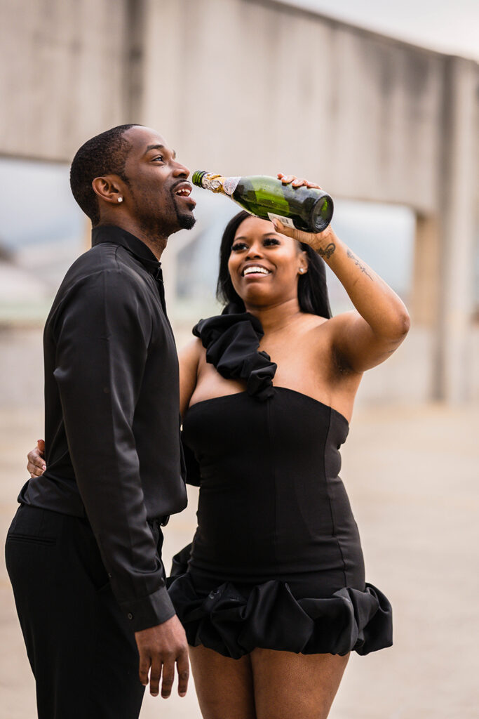 A Black woman goes to pour champagne out of the bottle into her fiancé's mouth for their parking garage engagement photos in Roanoke, Virginia.