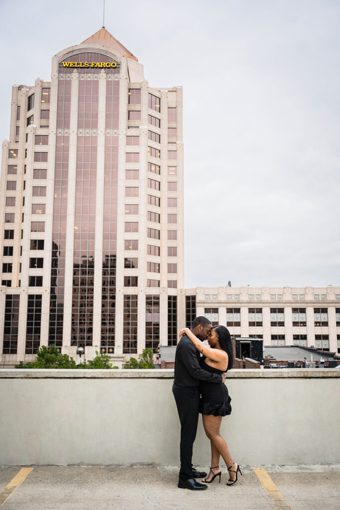 A Black couple goes in for a kiss at the top of a parking garage overlooking the iconic Wells Fargo tower in Roanoke, Virginia.