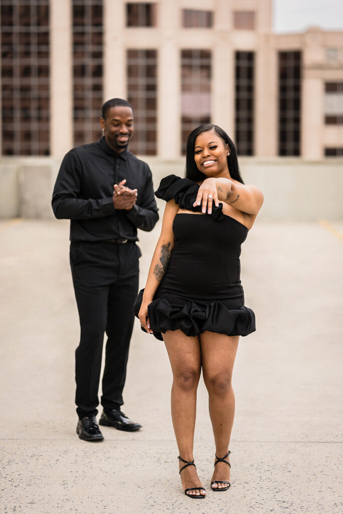A woman smiles and shows her engagement ring while her fiancé smiles proudly in the background.