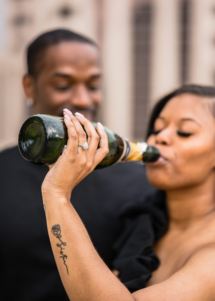 A Black woman drinks champagne directly from the bottle with her fiancé next to her during their engagement photoshoot.