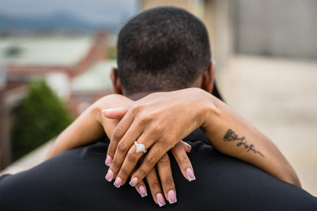 A woman wraps her arms around her fiancé's shoulders to show off her engagement ring for their parking garage engagement photos.
