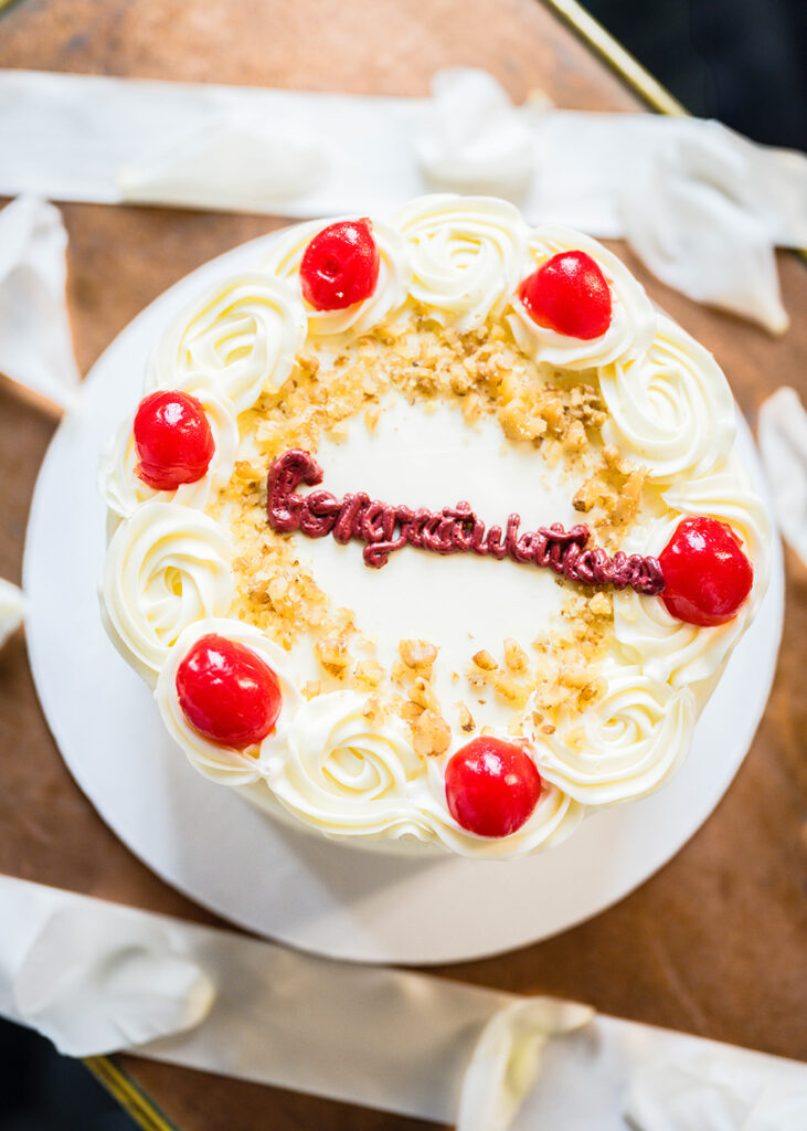 A small round decorated wedding cake is placed atop of a small table with flower petals and ribbons.