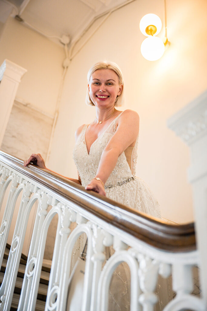 A bride holds onto the banister of a stairwell and smiles for a photo during her first look at the Liberty Trust Hotel.