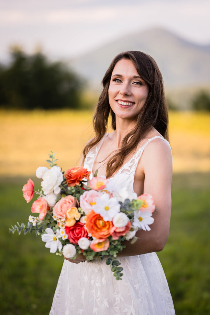 A woman wearing a white wedding dress smiles and holds her bouquet in front of her as she poses in front of Cahas Mountain golden hour Blue Ridge Parkway wedding portraits.
