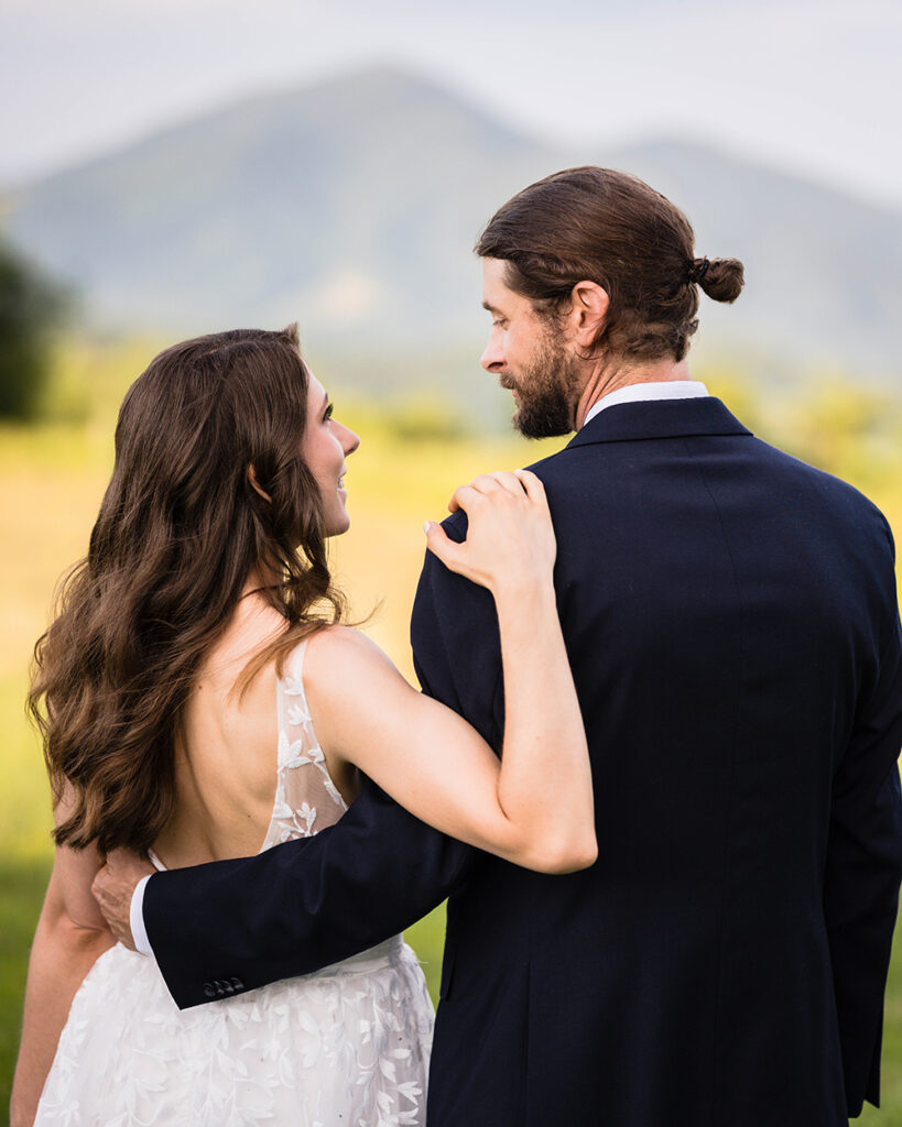 An elopement wedding couple stand together facing Cahas Mountain and steal a glance at one another during their golden hour portrait session.