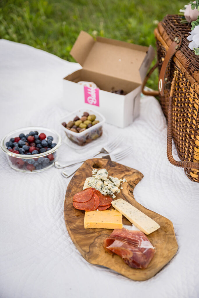 A close up shot of a charcuterie board with various meats and cheeses, a bowl of berries, a container of olives, a box with two cupcakes, and a picnic basket all placed atop of a white quilted blanket.
