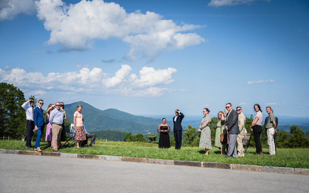 The groom, wedding officiant, and a group of wedding guests look as the bride and her Dad begin their walk down the aisle at Cahas Mountain Overlook.