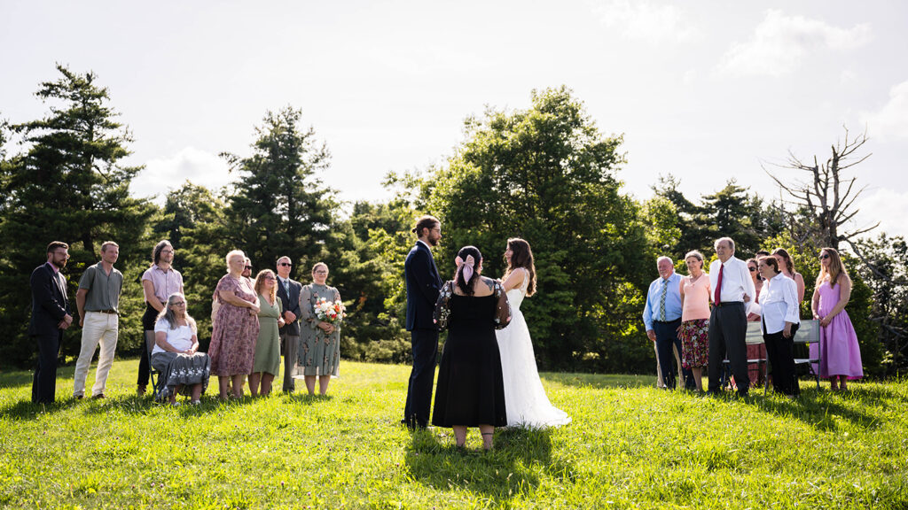 An elopement couple joins their officiant to begin their Blue Ridge Parkway elopement ceremony as their guests, in groups on either side of them, stand or sit as witness.